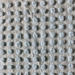 May include: Close-up of a light blue fabric with a textured surface. The fabric features a repeating pattern of raised, fluffy, and rounded tufts, creating a soft, tactile appearance. The background is a lighter shade of blue.