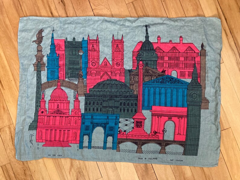 May include: A blue and pink linen tea towel with a design of famous London landmarks, including Big Ben, the Houses of Parliament, and St. Paul's Cathedral. The text "All Pure Linen" and "Made in Ireland" is printed on the towel.