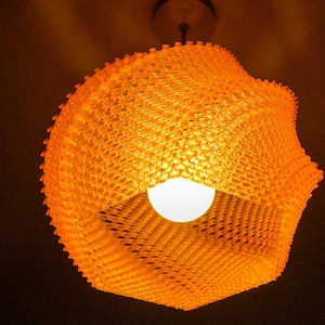 Könnte beinhalten: Eine orangefarbene, gewebte Pendelleuchte mit einer weißen Glühbirne. Das Licht hat ein einzigartiges, strukturiertes Design und hängt an einer Deckenleuchte. Die Leuchte hat eine warme orange Farbe.