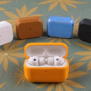 May include: A selection of colourful protective cases for wireless earbuds. The cases are in white, brown, blue, and black, with one open orange case revealing the earbuds. The earbuds are white.
