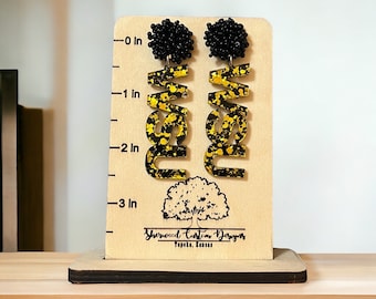 May include: A pair of black and gold statement earrings with a black bead accent. The earrings are shaped like the letters "NEW" and are approximately 3 inches long.