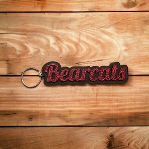 May include: A black keychain with a red glitter Bearcats word design. The keychain has a silver metal ring.