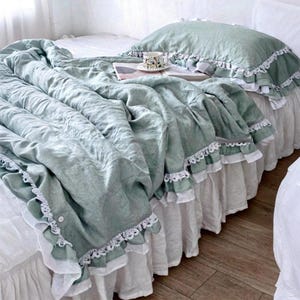 May include: A bed with a sage green comforter and pillow, both trimmed with white lace. The bed skirt is white with a ruffled design. A small teacup and saucer sit on a book on the bed.