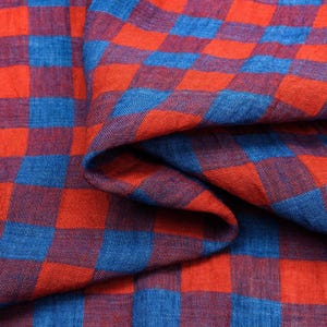 Può includere: Primo piano di un tessuto con un motivo a scacchi audace. Il design presenta quadrati alternati di rosso vibrante e blu intenso. Il tessuto è leggermente stropicciato, aggiungendo consistenza all'aspetto generale. Il materiale sembra morbido e leggero.