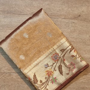 May include: A folded brown and cream colored silk saree with a floral pattern. The saree has a gold border with a brown trim.