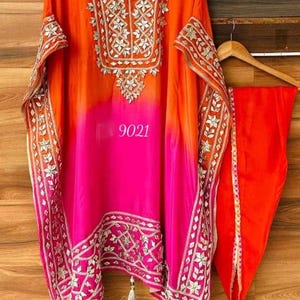 May include: A pink and orange ombre kaftan with silver embroidery and tassels. The kaftan has a V-neckline and is made of a lightweight fabric. The number 9021 is printed on the front of the kaftan.