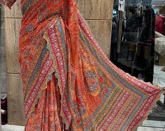 May include: An orange and pink saree with intricate embroidery and embellishments. The saree features a detailed border with blue, pink, and gold accents. The fabric drapes over a mannequin, showcasing the design and texture of the traditional Indian garment.