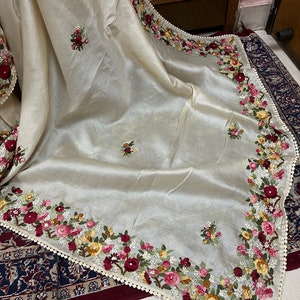 May include: A cream-colored silk shawl with delicate floral embroidery in shades of pink, red, yellow, and green. The shawl has a scalloped edge with a white lace trim.
