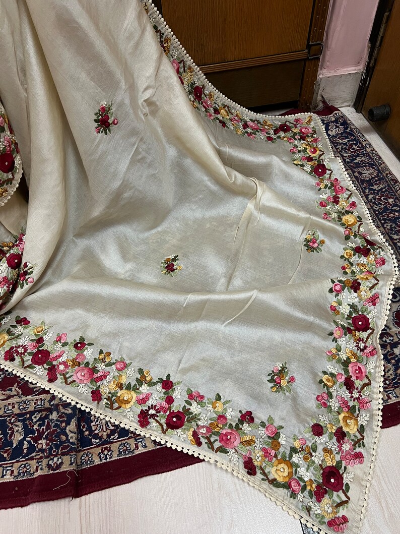 May include: A cream-colored silk shawl with delicate floral embroidery in shades of pink, red, yellow, and green. The shawl has a scalloped edge with a white lace trim.