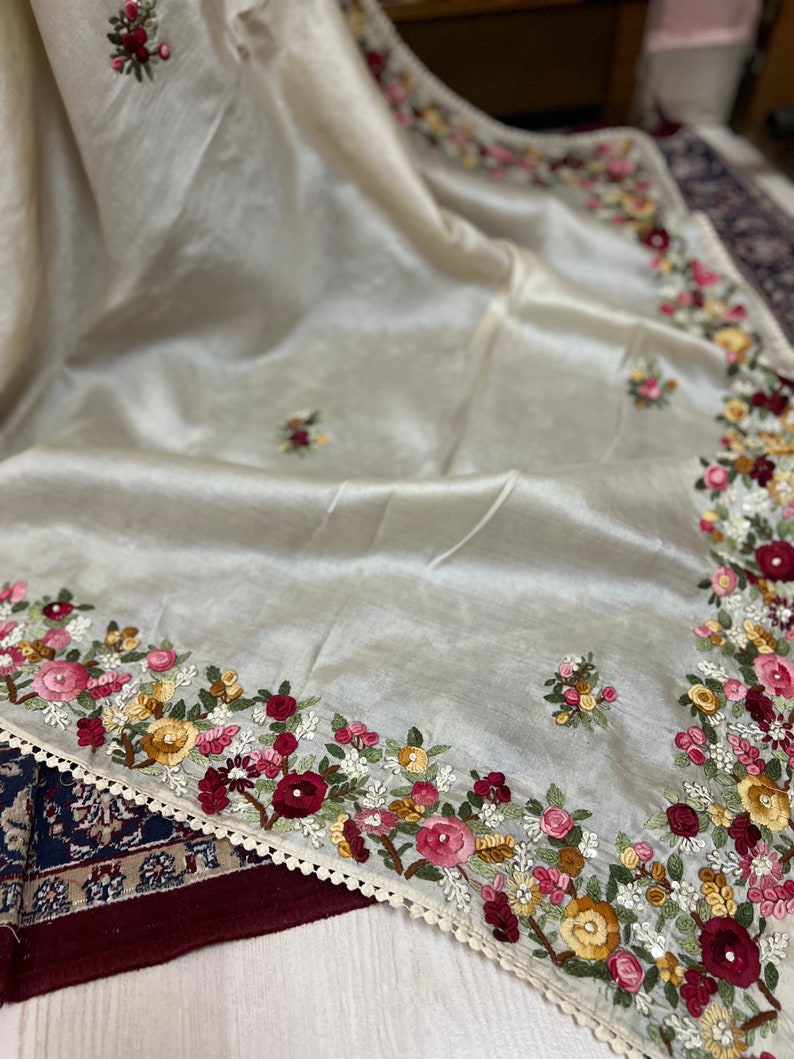 May include: A white silk scarf with a delicate floral embroidery design in shades of pink, red, yellow, and green. The embroidery is around the edges of the scarf and features a variety of flowers and leaves.