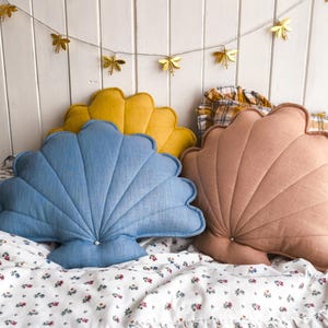 Może przedstawiać: Trzy poduszki w kształcie muszli w odcieniach niebieskiego, żółtego i brzoskwiniowego spoczywają na kwiecistej narzucie. Nad nimi wisi girlanda ze złotych światełek w kształcie ważek. Poduszki mają teksturowany, wachlarzowaty wzór.