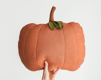 Puede incluir: Una mano sostiene una calabaza grande de tela naranja con una hoja de fieltro verde en la parte superior. La calabaza está hecha de una tela texturizada y tiene un aspecto ligeramente acolchado.
