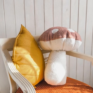 以下が含まれることがあります： 茶色のトップと白いボトムを持つ、キノコの形をしたぬいぐるみ枕。枕は白とオレンジのストライプの椅子の上に置かれています。