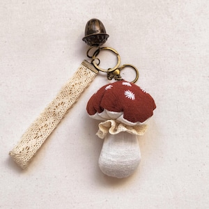 Op de afbeelding: Een handgemaakte paddenstoel sleutelhanger met een rode kap en witte geborduurde details. De sleutelhanger heeft een crèmekleurige kanten polsband en een bronzen eikelbedel. De paddenstoel heeft een witte gerimpelde kraag en een witte steel.