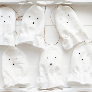 Op de afbeelding: Een set van zes witte stoffen spookornamenten, elk voorzien van twee geborduurde zwarte stippen als ogen en één zwarte stip als mond. Elk spookje is bevestigd aan een dun bruin touwtje om op te hangen en heeft een geplooide onderrand.