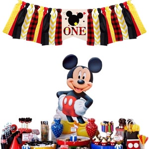 Puede incluir: Un conjunto de decoraci&oacute;n para una fiesta de primer cumplea&ntilde;os con tem&aacute;tica de Mickey Mouse. El conjunto incluye una pancarta con tela de cuadros rojos y negros, tela de chevron amarilla y blanca y tela negra con lunares amarillos. La pancarta tambi&eacute;n incluye una gran cabeza de Mickey Mouse con orejas negras y una cara blanca. La pancarta dice "ONE" en letras blancas. El conjunto tambi&eacute;n incluye un recorte de cart&oacute;n de Mickey Mouse, una mesa con varias decoraciones de fiesta y una variedad de golosinas.