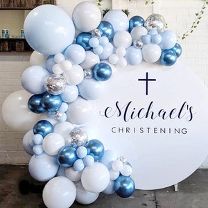 May include: A white round backdrop with a blue cross and the text "Michael's Christening" decorated with a garland of blue, white, and silver balloons.
