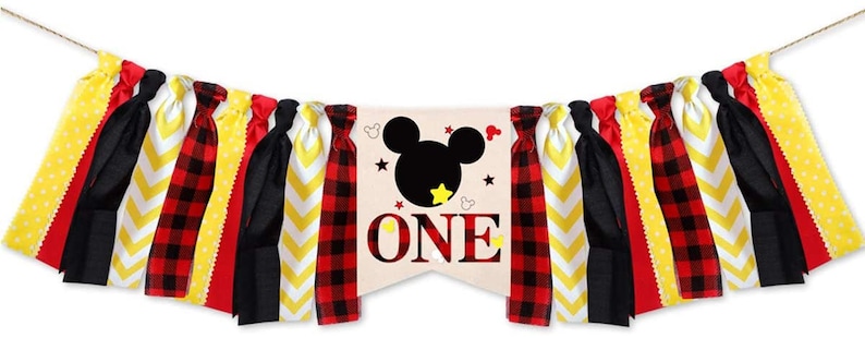 Puede incluir: Una pancarta de primer cumplea&ntilde;os con tem&aacute;tica de Mickey Mouse con tiras de tela roja, negra, amarilla y blanca. La pancarta presenta una cabeza de Mickey Mouse con una silueta negra y la palabra "ONE" en letras rojas.