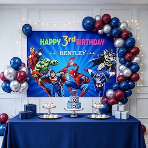 May include: A birthday party backdrop featuring superhero illustrations, the text HAPPY 3rd BIRTHDAY BENTLEY, and a balloon arch in blue, silver, and red. A two-tier cake and cupcakes are on a blue tablecloth with gift boxes on the sides.