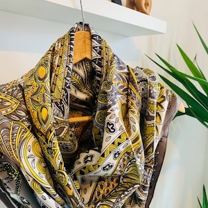 May include: A brown and yellow patterned silk scarf with a paisley design. The scarf is hanging on a hanger.