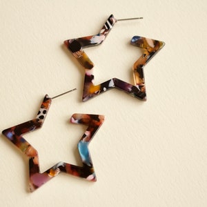 May include: A pair of star-shaped earrings with a multi-coloured, abstract pattern. The earrings are made of a translucent material and have a silver post.