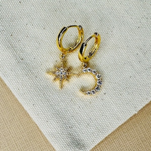 May include: A pair of gold hoop earrings with a dangling star and crescent moon charm. Both charms are adorned with clear cubic zirconia stones.