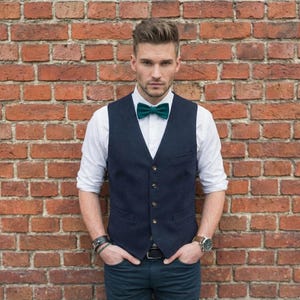May include: A man's formal attire, featuring a navy blue vest with gold buttons, a white dress shirt, and a teal bow tie. The outfit is completed with dark blue jeans and a watch. The background is a red brick wall.
