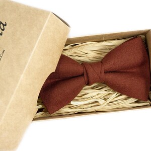 Puede incluir: Una pajarita de color óxido descansa dentro de una caja de cartón llena de paja. La caja está parcialmente abierta, revelando la pajarita. La caja tiene el texto "Urba Vana" impreso.