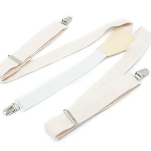 Puede incluir: Tirantes blanquecinos con clips metálicos plateados. Una correa es blanca y la pieza central es de color beige claro. Los tirantes están colocados sobre un fondo blanco.