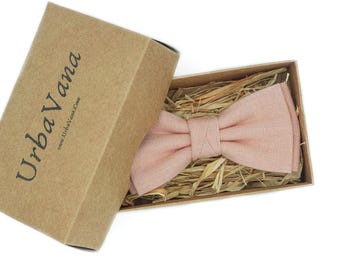 May include: A light pink fabric bow tie rests inside a rectangular brown cardboard box filled with straw. The box lid is open and features the black printed text UrbaVana and the website address www.UrbaVana.com on the exterior side.