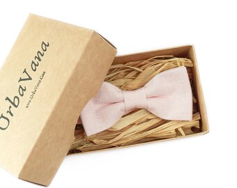May include: A light pink bow tie rests inside a rectangular cardboard box filled with shredded paper filler. The box lid is propped open and features the black printed text UrbaVana and the website address www.UrbaVana.com on the side.