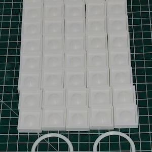 Puede incluir: Un conjunto de 48 cuadrados de plástico blanco con una cúpula elevada en el centro. Los cuadrados están dispuestos en una cuadrícula. Dos anillos de plástico blanco se encuentran debajo de los cuadrados.