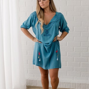 May include: A woman wearing a light blue, embroidered, short-sleeved dress with tassels. The dress has a V-neck and a loose fit.