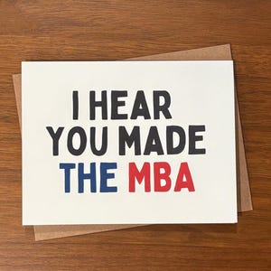May include: A white card with the message "I HEAR YOU MADE THE MBA" in black, blue, and red lettering. The card sits on a brown envelope, against a wooden background. A congratulatory card.