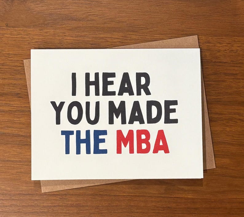 May include: A white card with the message "I HEAR YOU MADE THE MBA" in black, blue, and red lettering. The card sits on a brown envelope, against a wooden background. A congratulatory card.