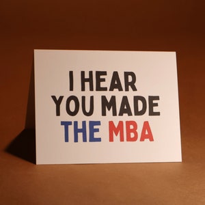 May include: A white card with the message "I HEAR YOU MADE THE MBA" in black, blue, and red. The card is propped up against a brown backdrop. The text is in a bold, handwritten font.