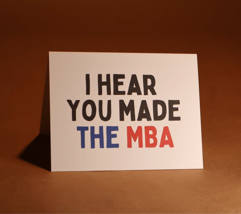 May include: A white card with the message "I HEAR YOU MADE THE MBA" in black, blue, and red. The card is propped up against a brown backdrop. The text is in a bold, handwritten font.