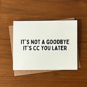Pu&ograve; includere: Un biglietto di auguri bianco con il testo "IT'S NOT A GOODBYE IT'S CC YOU LATER" in nero, appoggiato su una busta marrone. Il biglietto e la busta sono su una superficie di legno.
