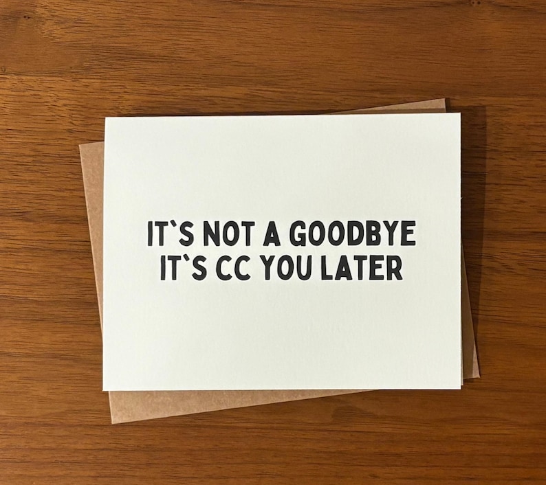 Pu&ograve; includere: Un biglietto di auguri bianco con il testo "IT'S NOT A GOODBYE IT'S CC YOU LATER" in nero, appoggiato su una busta marrone. Il biglietto e la busta sono su una superficie di legno.