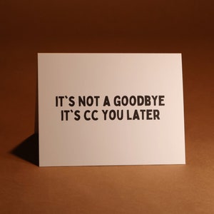 Pu&ograve; includere: Un biglietto bianco con testo nero: "IT'S NOT A GOODBYE IT'S CC YOU LATER". Il biglietto rettangolare &egrave; posto su uno sfondo marrone.