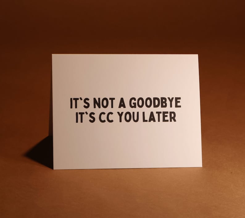 Pu&ograve; includere: Un biglietto bianco con testo nero: "IT'S NOT A GOODBYE IT'S CC YOU LATER". Il biglietto rettangolare &egrave; posto su uno sfondo marrone.