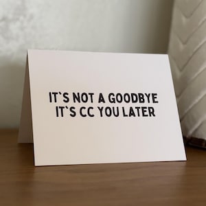 Pu&ograve; includere: Un biglietto di auguri bianco con testo nero che recita "IT'S NOT A GOODBYE IT'S CC YOU LATER". Il biglietto &egrave; piegato e appoggiato su una superficie di legno. Un cuscino decorativo &egrave; sullo sfondo.