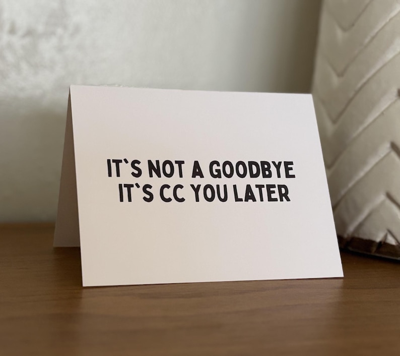 Pu&ograve; includere: Un biglietto di auguri bianco con testo nero che recita "IT'S NOT A GOODBYE IT'S CC YOU LATER". Il biglietto &egrave; piegato e appoggiato su una superficie di legno. Un cuscino decorativo &egrave; sullo sfondo.