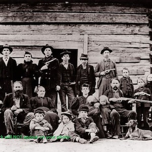 Pode incluir: Uma fotografia a preto e branco de um grande grupo familiar em pé em frente a uma cabana de madeira. A família está vestida com roupas do início dos anos 1900. Alguns membros da família estão segurando armas de fogo.