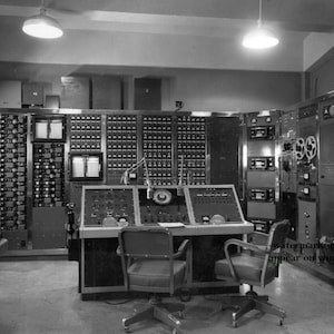Peut inclure: Une photo en noir et blanc d'une salle de contrôle informatique vintage avec un grand panneau de commutateurs et de cadrans, deux chaises et un bureau avec un microphone.
