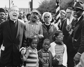 May include: A black and white photograph of a group of people protesting. A man in a suit and clerical collar leads the group, with a woman in a white hat and a man in a hat and sunglasses behind him. The group is holding signs and chanting.