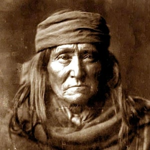 May include: A black and white portrait of a Native American man wearing a brown headband and a brown blanket. He has long dark hair and a serious expression.