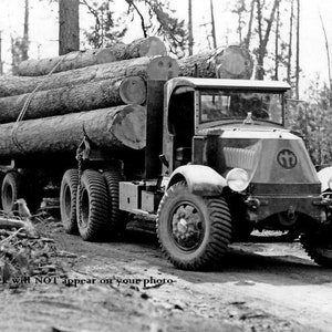 May include: A black and white photo of a large truck hauling a load of logs. The truck has a large, round grill with a logo in the center. The truck is driving on a dirt road.