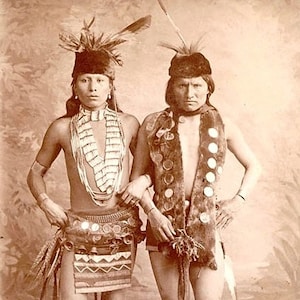 May include: Two Native American men wearing traditional clothing and headdresses. The man on the left is wearing a beaded necklace and a skirt with bells. The man on the right is wearing a fur vest with metal accents.