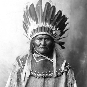 May include: Black and white portrait of a Native American man wearing a traditional headdress with feathers. He is wearing a fringed leather garment and has a serious expression.